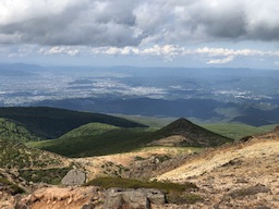 a picture of the beautiful view on top of Mt. Adatara in Fukushima, Japan. In the distance is a small valley of gusting green and arching waves of city landscapes.